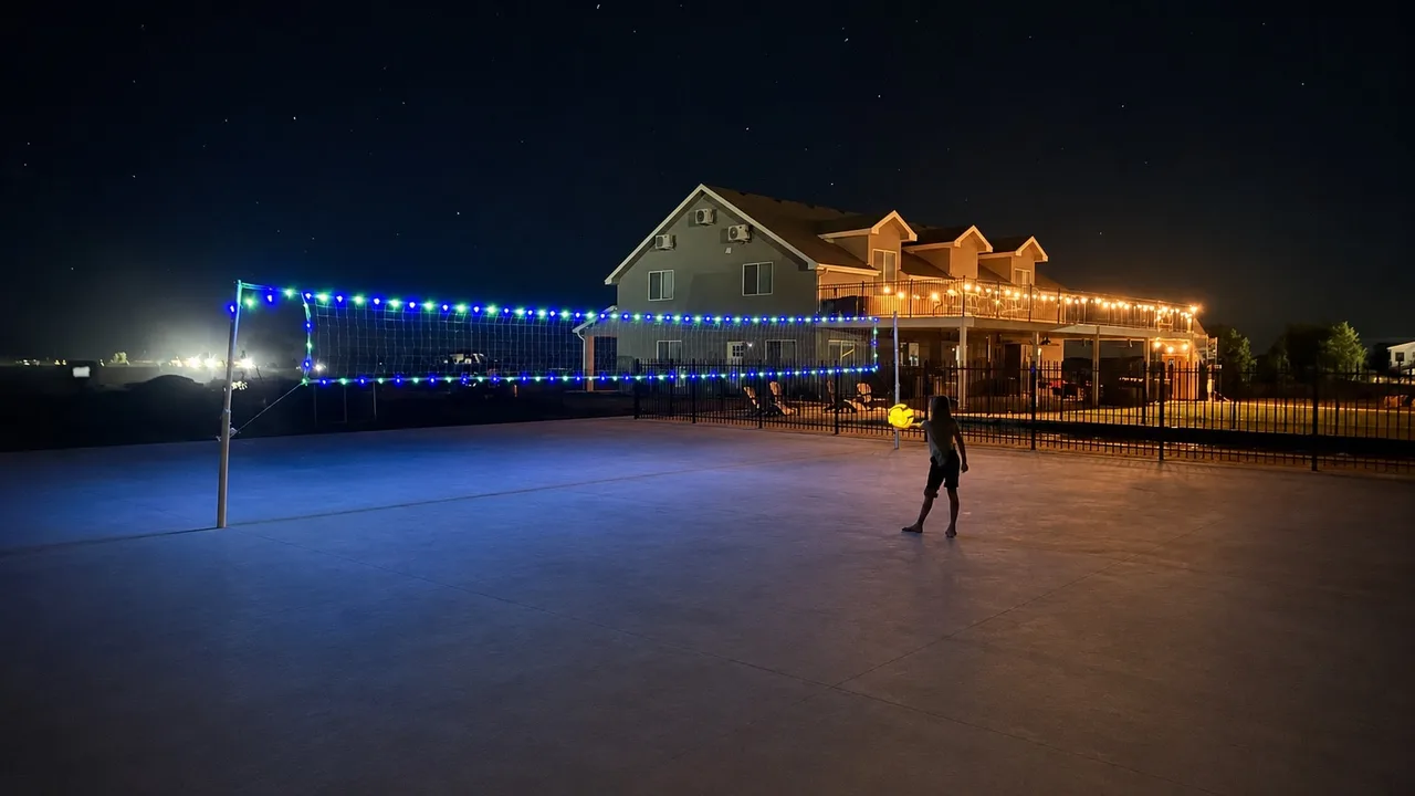Pickleball court at night lit by blue and green LED string lights along the net, with a player mid-serve and the retreat's warm windows glowing behind.